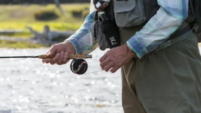 Close-up of hands holding a fly-fishing rod and managing the line beside a river.
